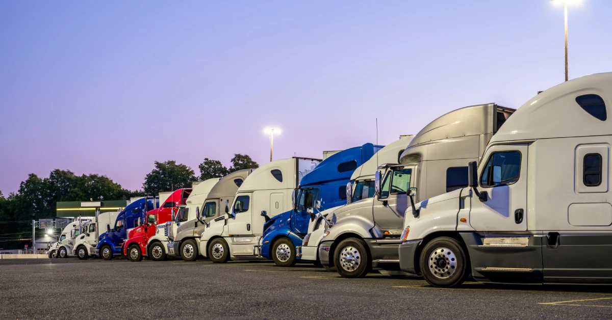 A row of semi-trucks with various color cabs parking in a semi-truck parking lot with street lights in the background.