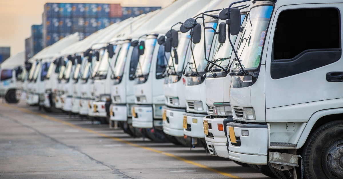 A long line of white semi trucks parked next to each other. There are some cargo containers stacked up behind them.