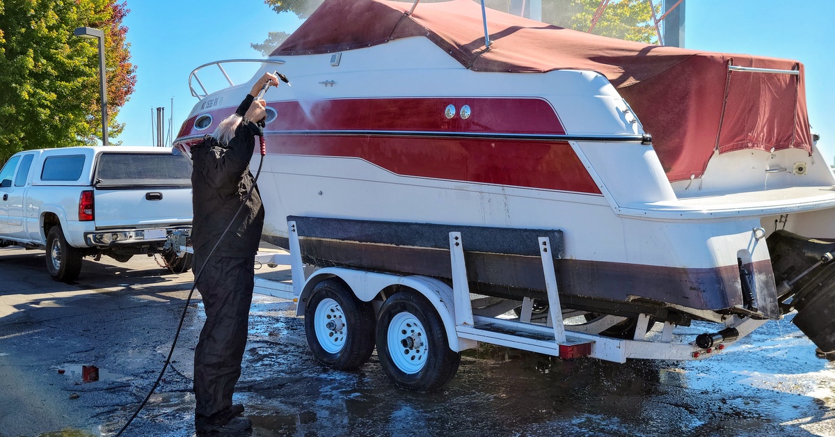 A man using a pressure washer to clean his boat. The boat is on a trailer and has a red cover over the top of it.