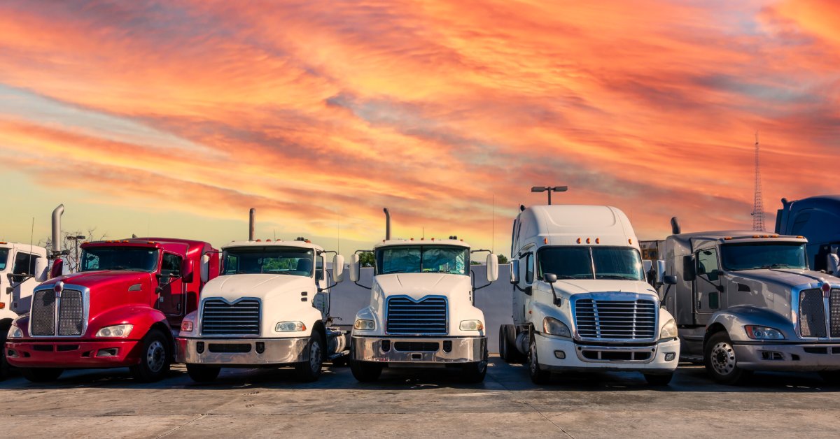 A row of semi truck cabs parked side by side in a paved lot under an orange sunset sky near a large building.