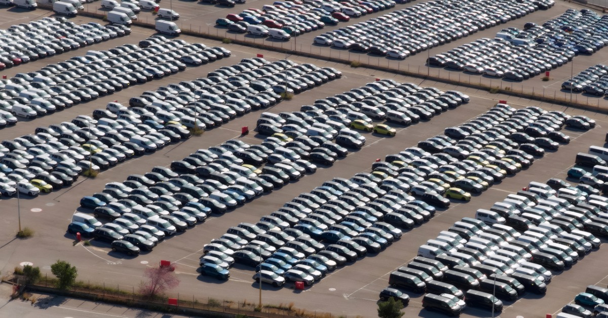 An aerial view of cars parked in precise rows across a large outdoor lot surrounded by fences, forming a dense pattern.