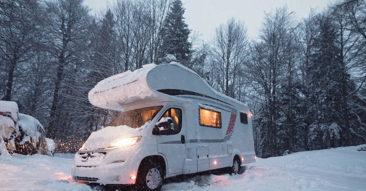 An RV sits outside in nature during the winter season. Snow covers the vehicle, the ground, and the trees.