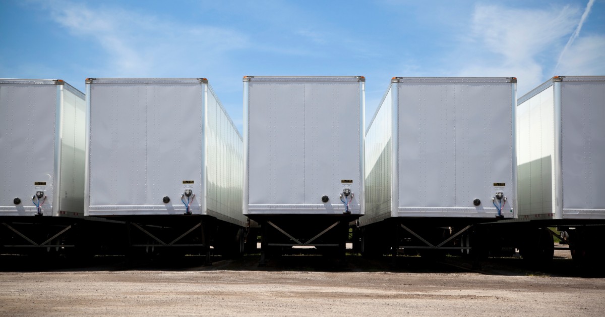 Five transport trucks sitting in an outdoor long-term storage facility, framed by a gravel road below and clear blue skies above.
