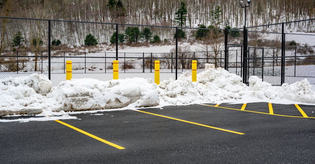 A freshly plowed parking lot with yellow-lined spaces, snow piled along the fence, and a wintery hillside in the background.