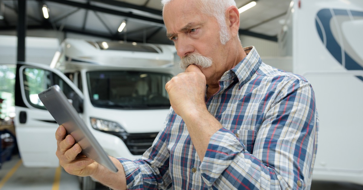 A man in a flannel shirt touches his chin while looking at an electronic tablet with multiple motorhomes and RVs behind him.