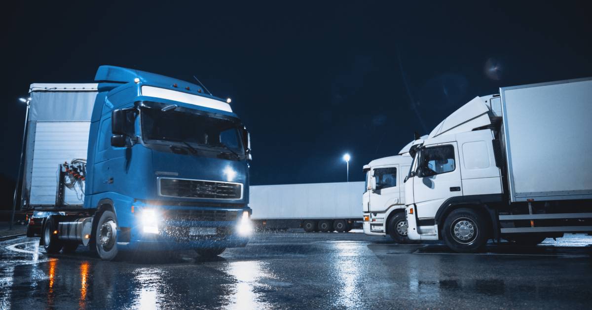 A truck with a blue cab drives into a parking lot filled with other semi-trucks at night while it’s raining.