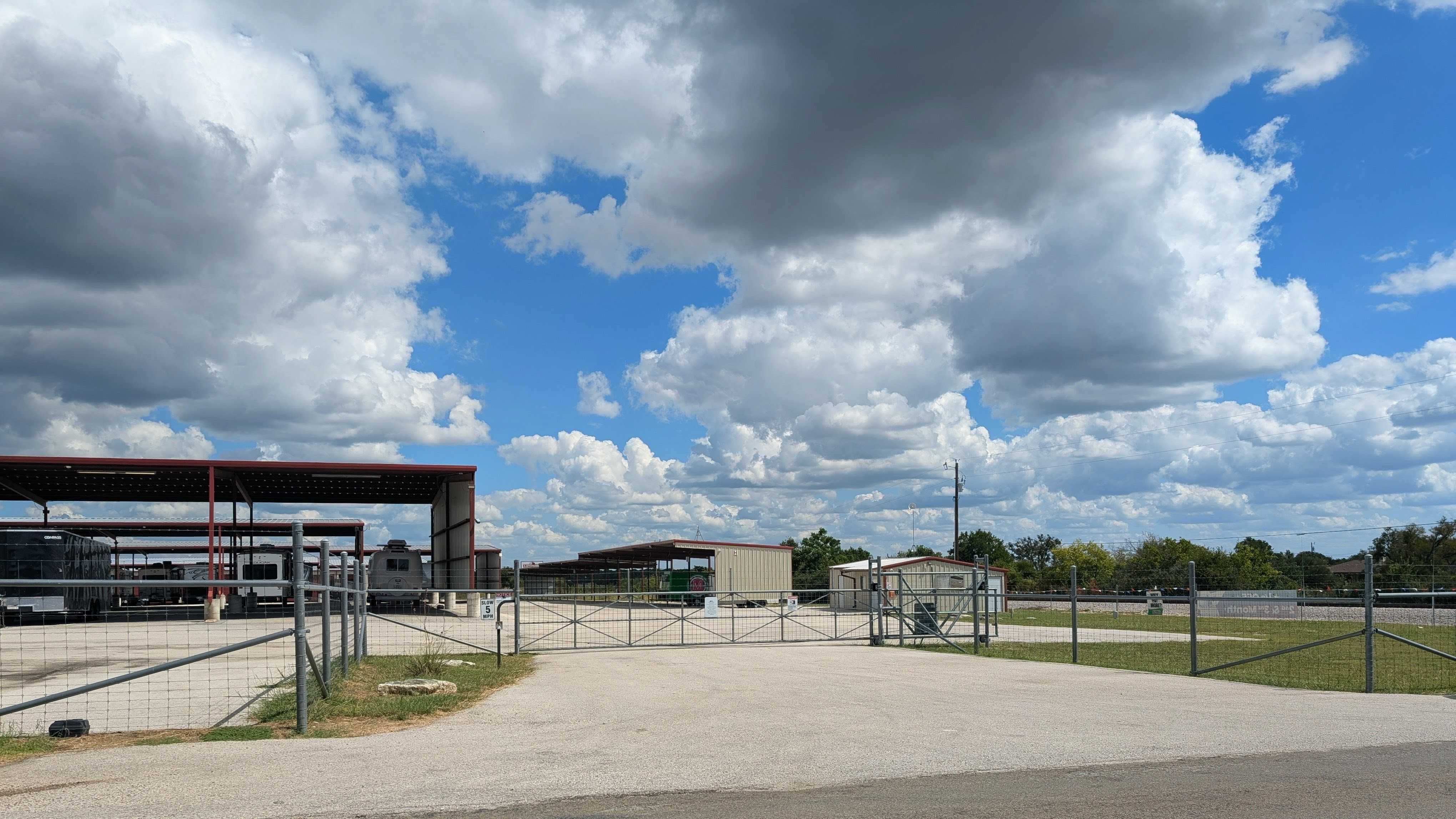 Secured gate entrance to an outdoor storage facility with covered parking stalls for RVs and boats under a bright, cloudy sky.