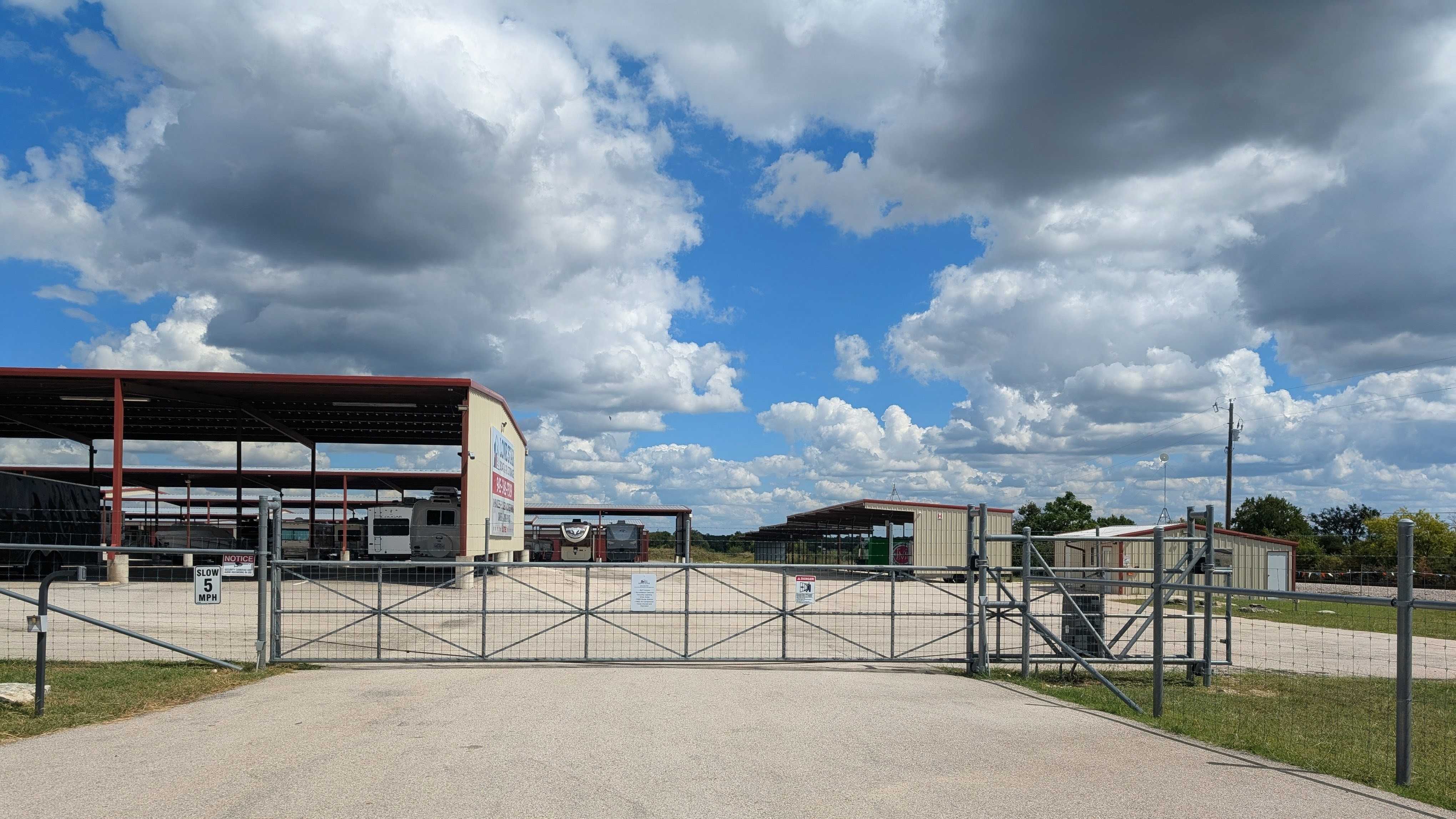 Wide metal security gate at the entrance to a facility with rows of covered RV and boat storage stalls under a dramatic cloudy sky.