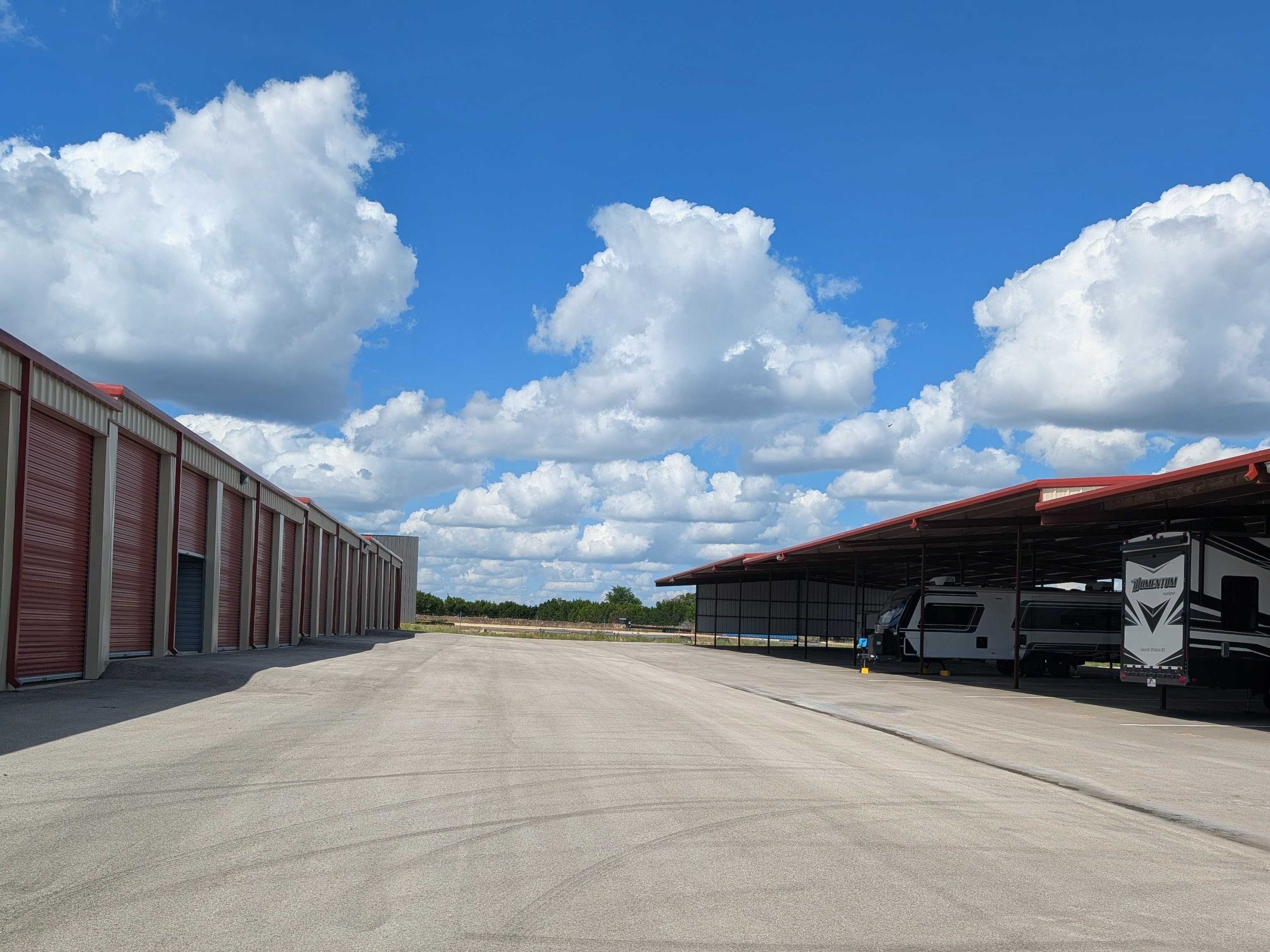 Wide concrete drive aisle between a row of enclosed storage units with red roll-up doors and a row of covered parking stalls holding RVs and boats.