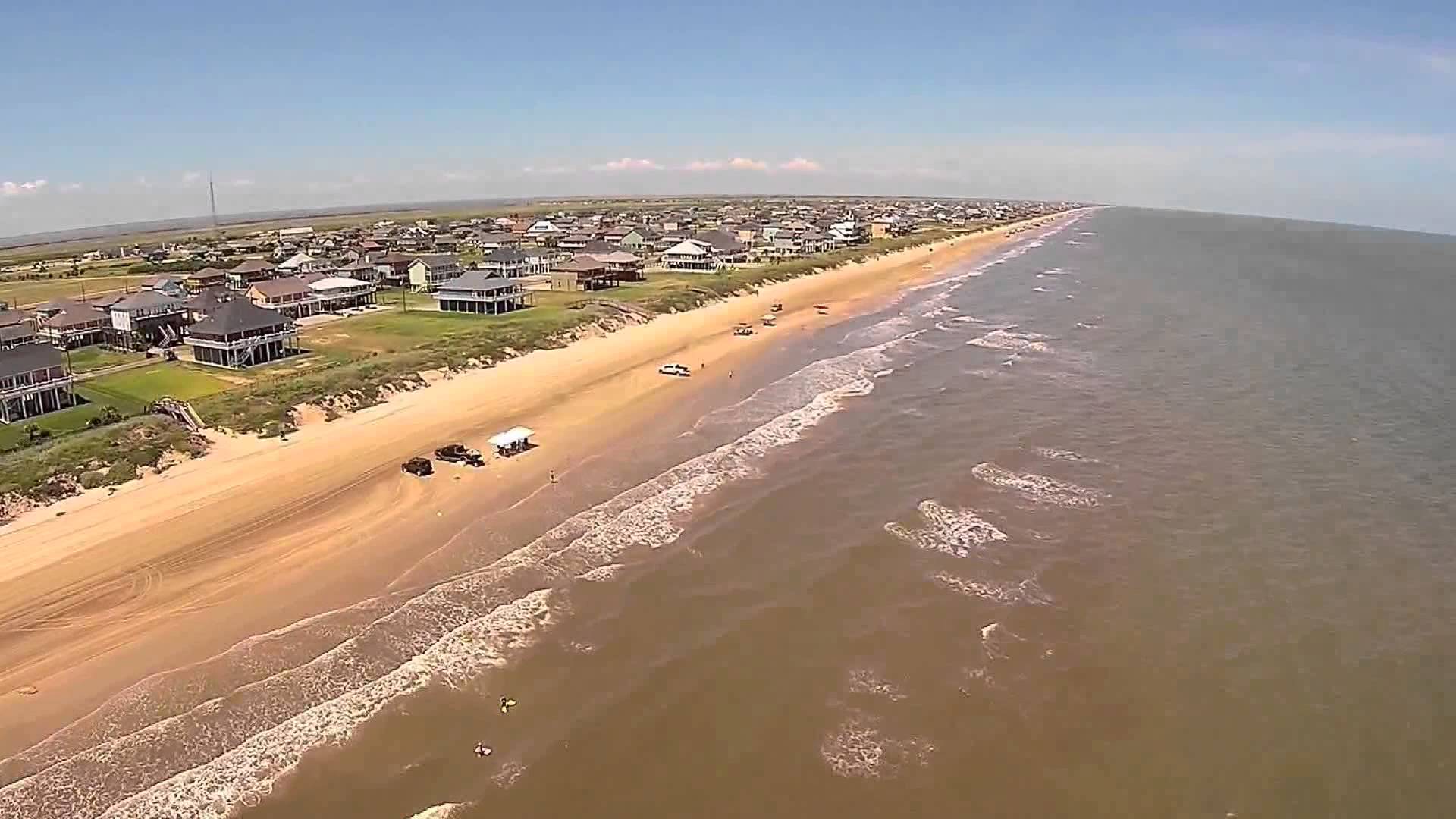 An expansive aerial view of the sandy coastline at Crystal Beach on the Bolivar Peninsula, featuring beachfront houses, driving-accessible shorelines with parked vehicles, and the gentle waves of the Gulf of Mexico under a clear blue sky. Aerial View of Crystal Beach and Port Bolivar, Texas