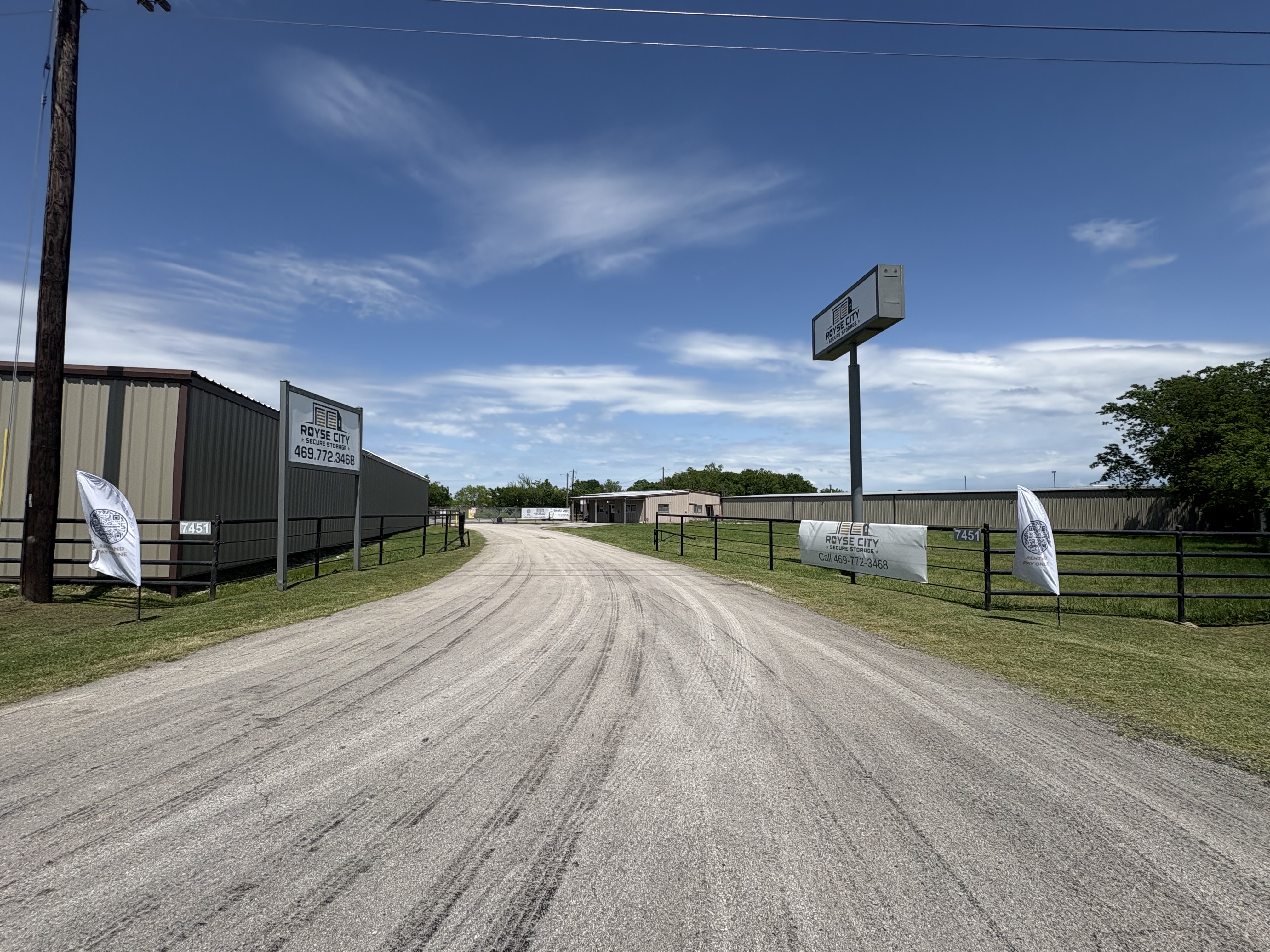 Entrance view of Royse City Secure Storage facility at 7451 FM 35, Royse City, TX, featuring gated access, signage with contact number (469-772-3468), and surrounding metal buildings under a clear blue sky.