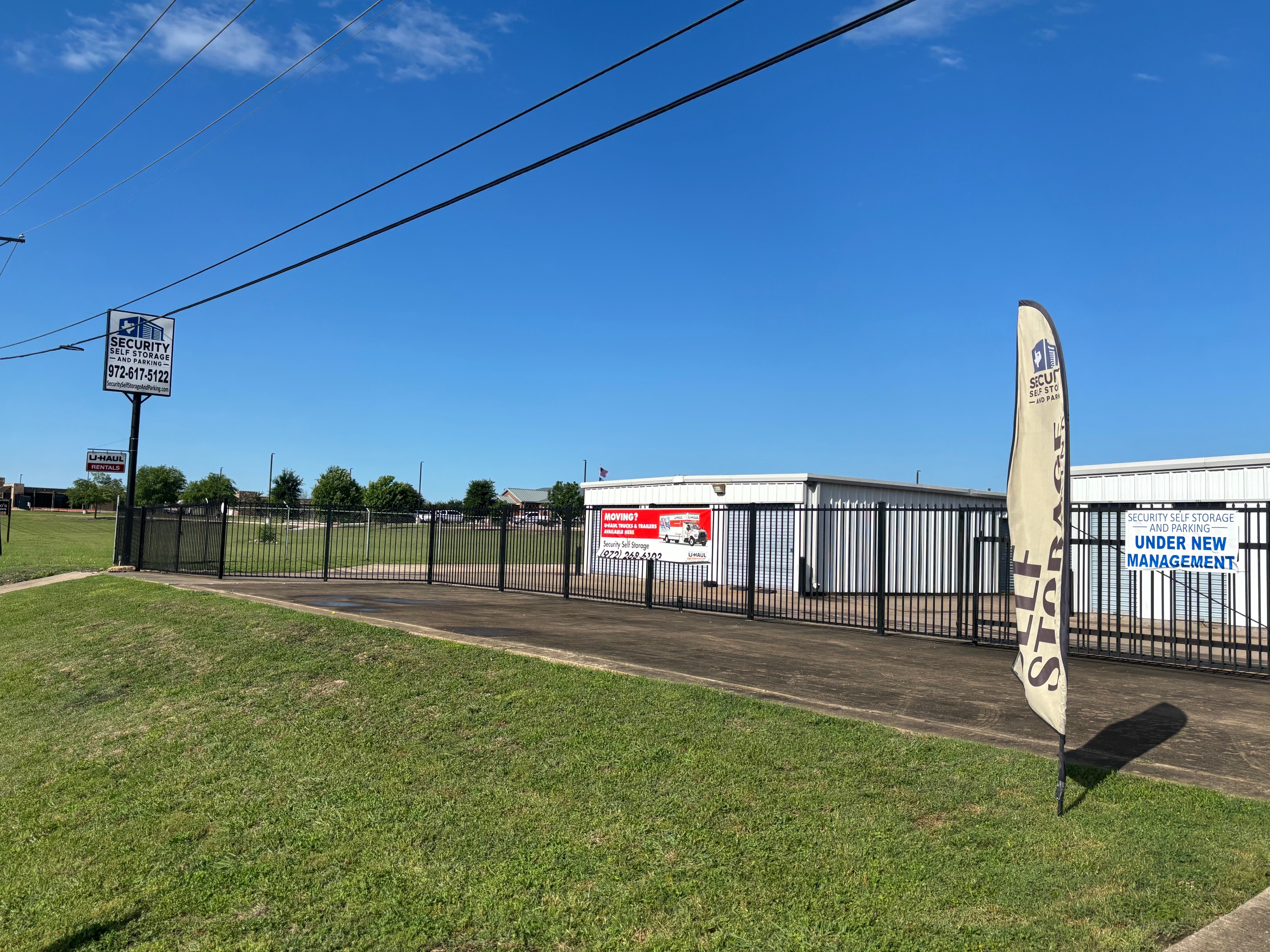 Security Self Storage and Parking facility entrance in Waxahachie, TX with gated access and on-site U-Haul rentals Gated entrance of Security Self Storage and Parking in Waxahachie, TX with signage, flag, and U-Haul rental banner under a clear blue sky.