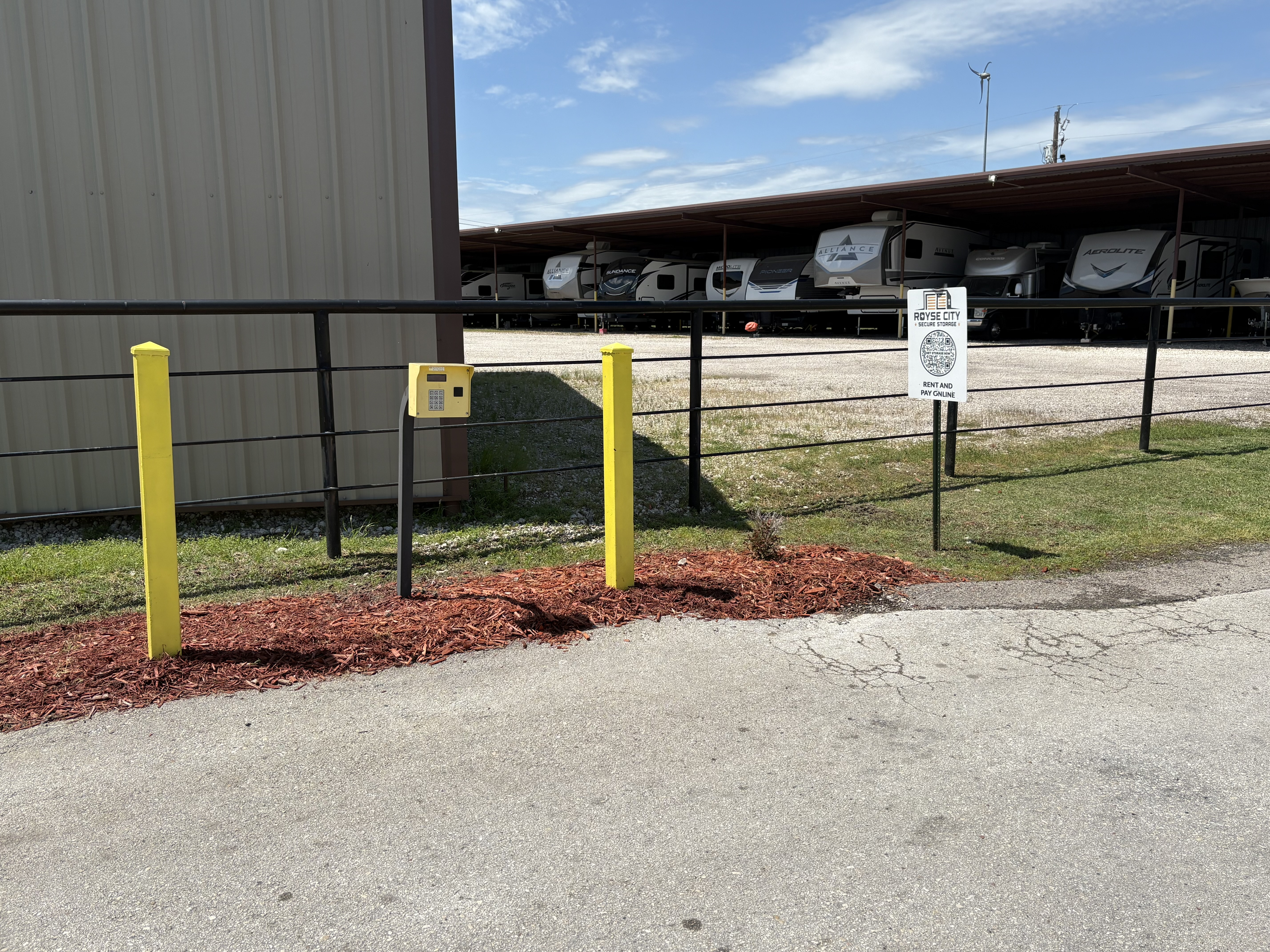 Royse City Secure Storage gated keypad entry with yellow safety posts, adjacent to covered RV parking area and online rental signage