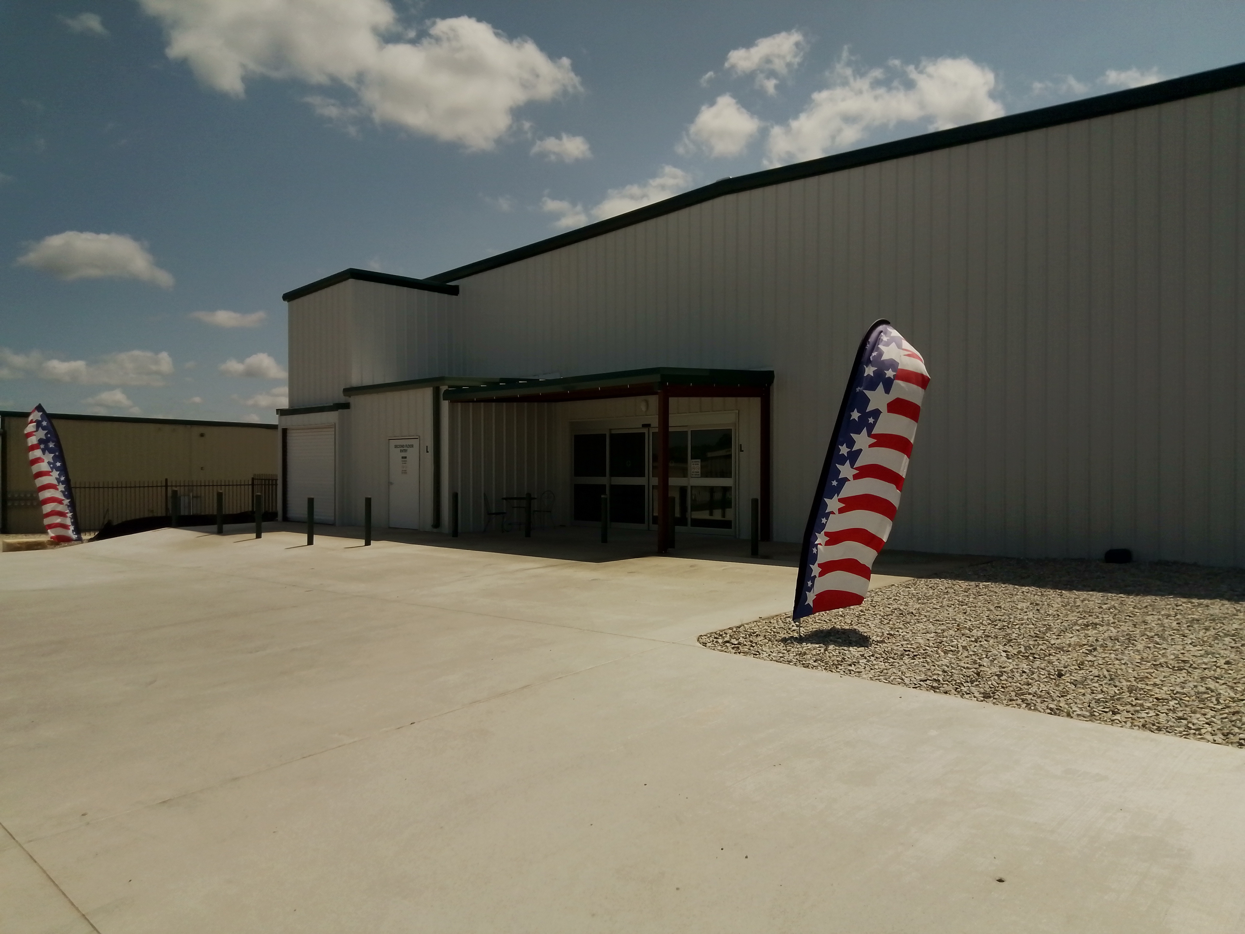 Exterior of a modern self-storage facility with American flag banners, wide concrete driveway, and metal siding under a sunny sky in Spicewood, Texas