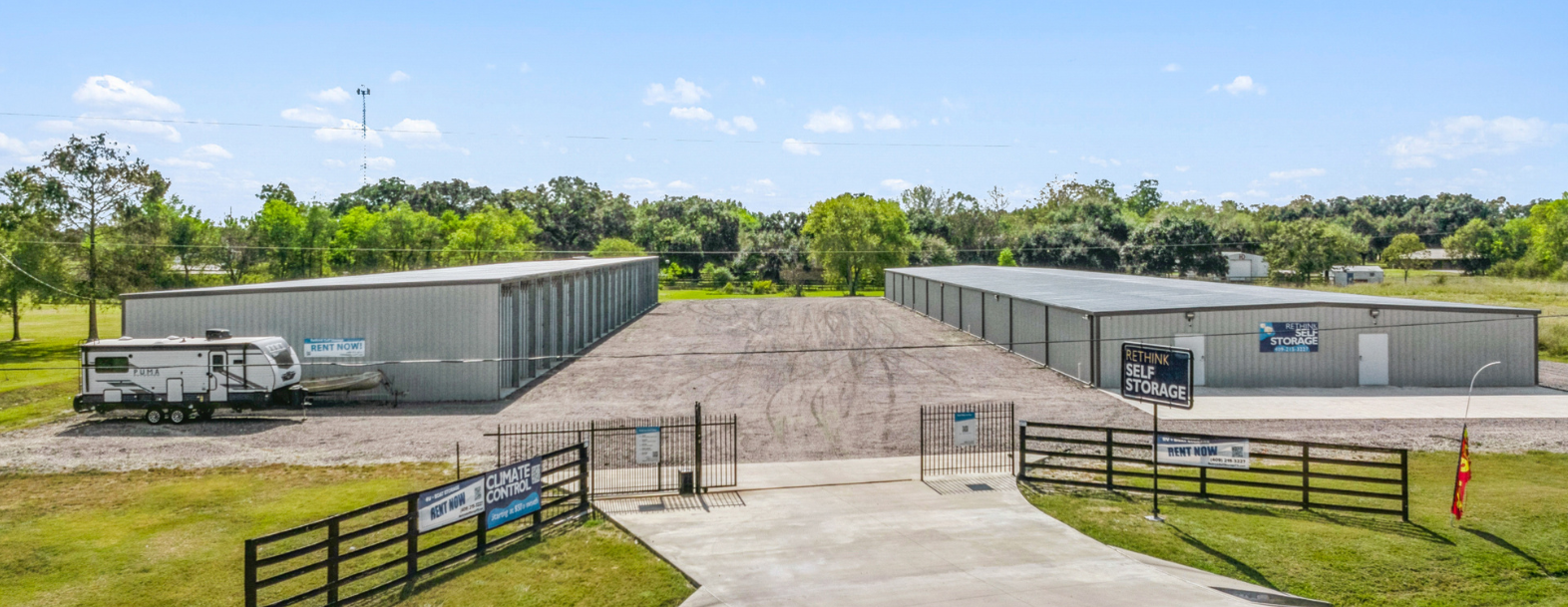 Wide view of Rethink Self Storage in Hamshire, TX, showing two large climate-controlled storage buildings, secure gated access, and outdoor parking space for RV and boat storage. The facility is surrounded by green trees and open space.