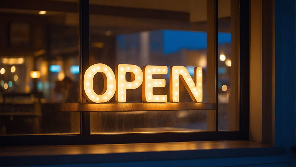 This image showcases a warmly lit "OPEN" sign, glowing brightly through a window. The sign is composed of large, bulb-lit letters, creating a welcoming and inviting atmosphere. The background features a dimly lit interior, likely a café or a small business, with a cozy ambiance. The warm lighting inside contrasts with the cool tones of the evening outside, adding to the overall charm and appeal of the scene. It captures a moment of readiness to welcome guests, indicating that the place is open for business.