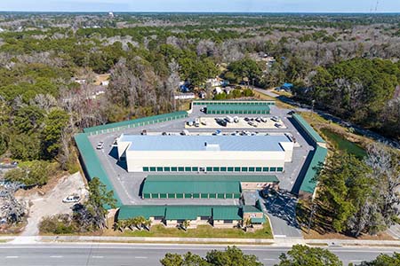 High-angle aerial view of a large, fenced self-storage facility surrounded by dense trees, featuring green-roofed buildings and open vehicle parking.