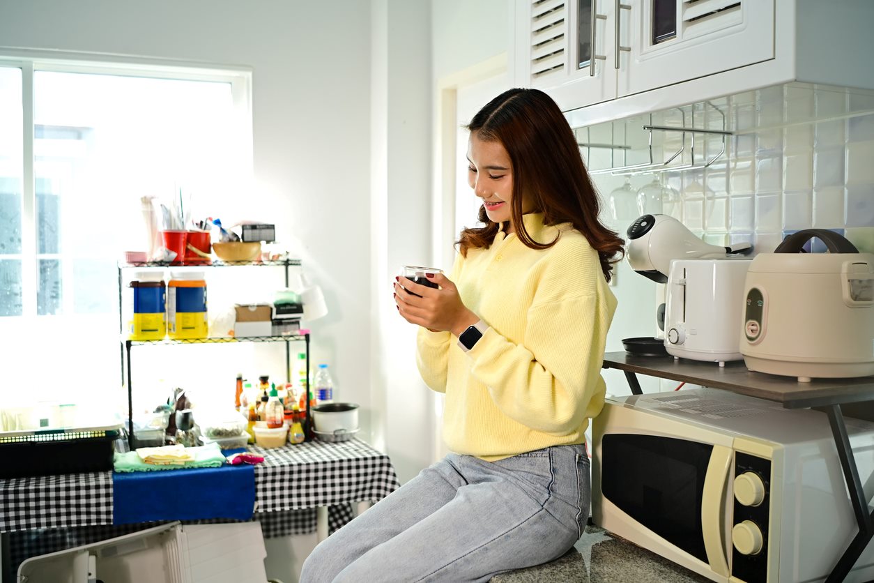 A young woman happily sits on her kitchen counter with her morning coffee in hand. 