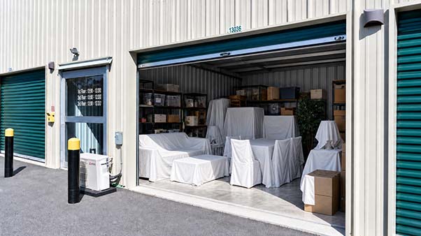 An open self-storage unit neatly packed with sheet-covered furniture, shelves, and cardboard boxes.