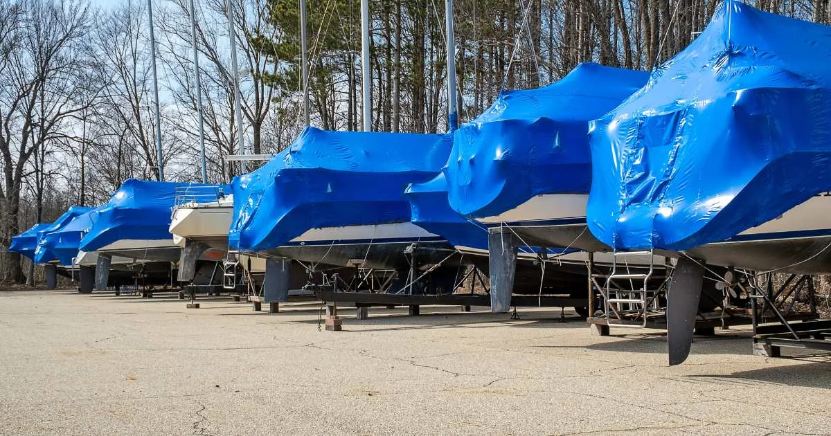 A row of sailboats covered in blue wrap, lined up in outdoor storage during off-season.