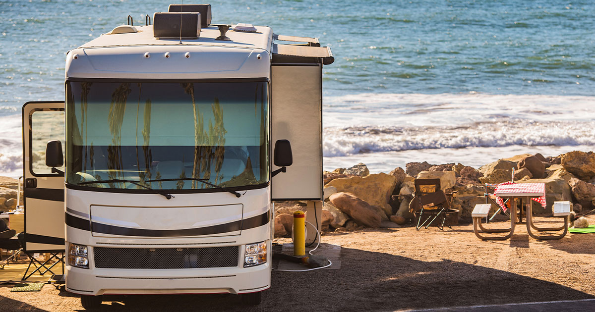 A modern RV parked near a scenic oceanfront with waves crashing against rocks, a picnic table with a red-checkered tablecloth, and camping chairs set up on the sandy ground.