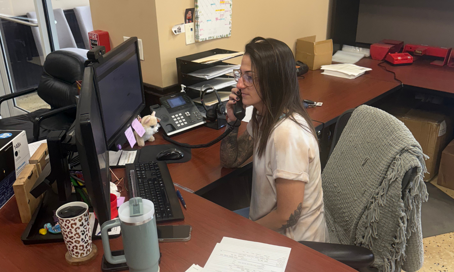 A woman seated at a desk in an office, speaking on the phone while working on a computer. The desk is organized with two monitors, paperwork, a headset, and a couple of drink tumblers. A small stuffed dog and notes are attached to one of the monitors. The atmosphere suggests a busy yet comfortable workspace.