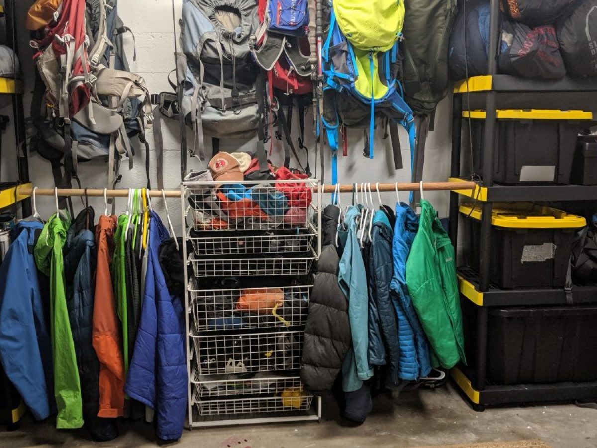 A neatly organized storage area featuring colorful jackets hanging on a rod, hiking backpacks hung on the wall, and sturdy black-and-yellow plastic bins stacked beside wire shelves — showcasing efficient gear storage and organization in a self-storage unit. Maximize Your Space: Smart Storage Solutions for Outdoor Gear and Seasonal Essentials
