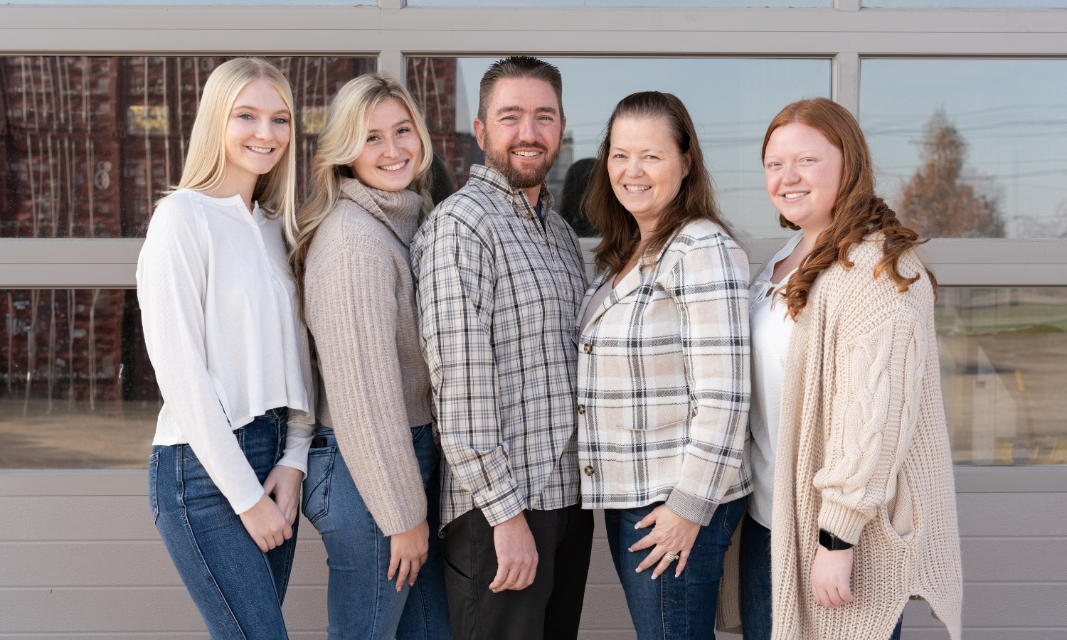 The Verdi family posing together in front of a glass-paneled building. Parents Joe and Patty stand at the center, with their daughters Kloe, Kourtney, and Rose standing from left to right. All are smiling and dressed in casual, cozy clothing.