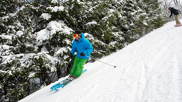 person in blue ski jacket and green pants skiing down a snowy slope