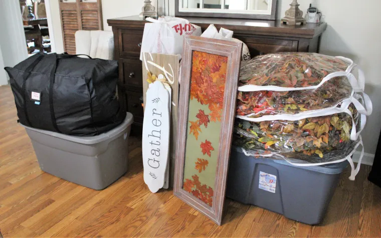 Plastic storage bins, a large black bag, and seasonal fall decorations—including a framed autumn leaf print, wreaths in clear bags, and a “Gather” sign—are stacked neatly on a hardwood floor in a living room.