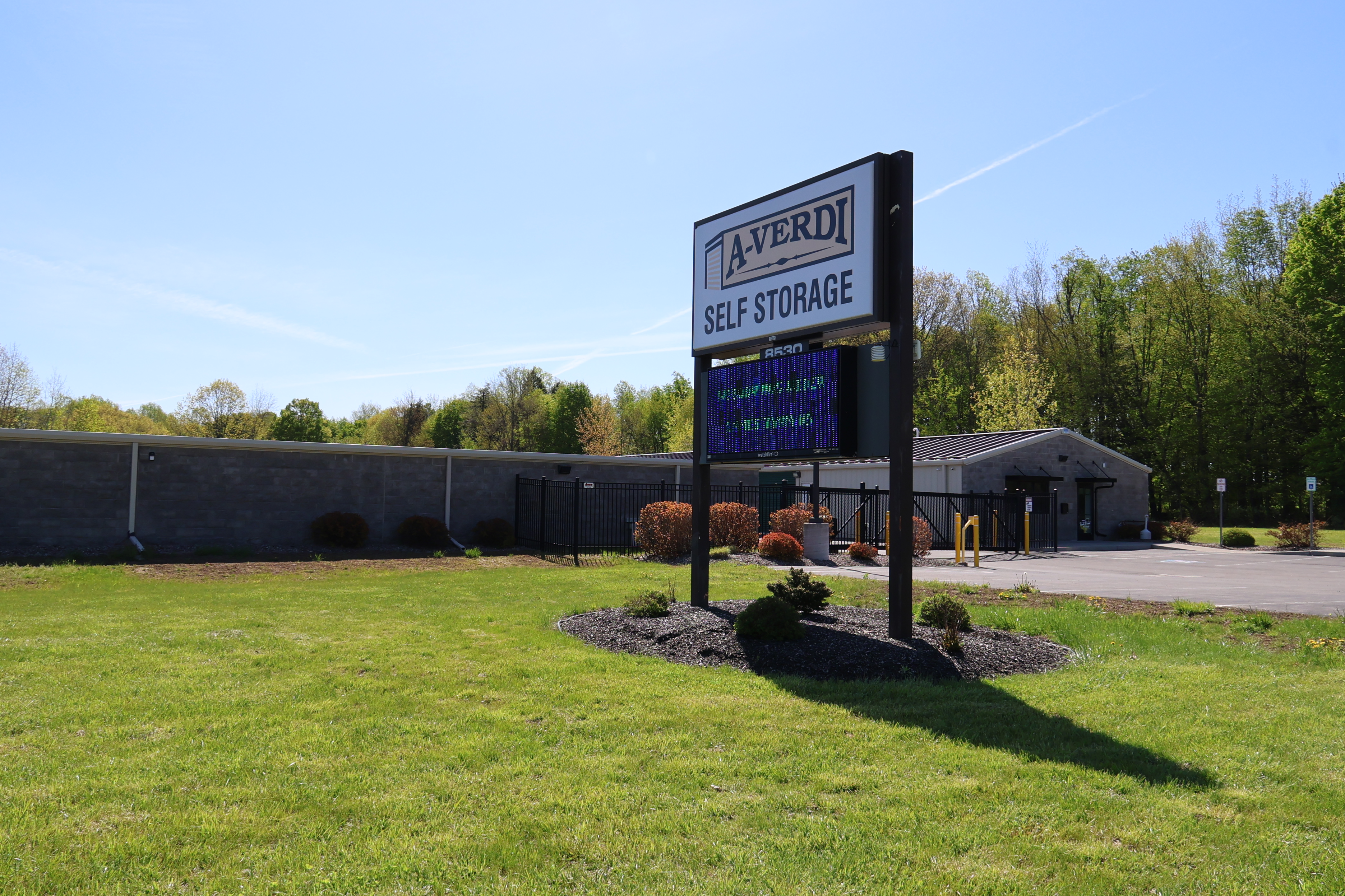 Exterior view of an A-Verdi Self Storage facility on a sunny day, showing the roadside sign, gated entrance, storage building, parking area, grass, and trees in the background.