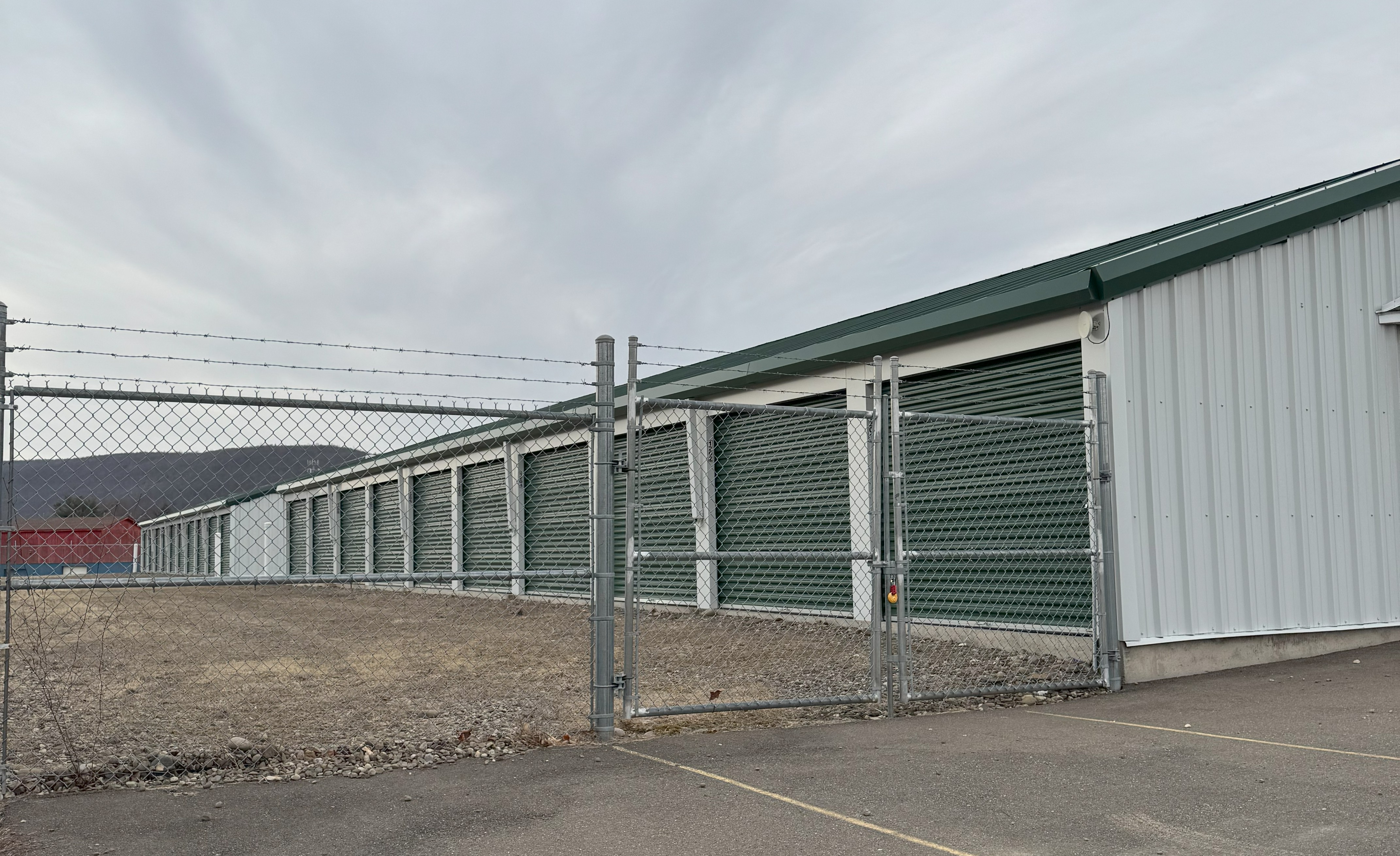 Row of secure self-storage units with green roll-up doors, enclosed by a tall chain-link fence topped with barbed wire. The storage facility sits on a paved lot with mountains in the background under a cloudy sky.
