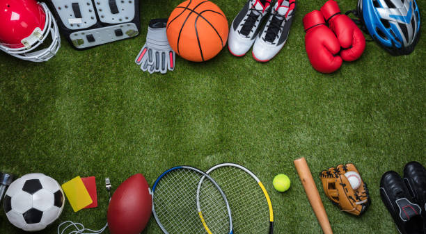 Various sports equipment on green turf including football helmet, soccer ball, basketball, tennis rackets, boxing gloves, baseball gear, and cycling helmet.