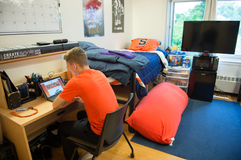 A college student sits at a wooden desk in a neatly organized dorm room, working on a laptop. The room has a single bed with blue bedding and an orange Syracuse pillow, a bean bag chair, a TV on top of a mini fridge, and plastic drawers filled with snacks. Posters decorate the walls, and school supplies are arranged on the desk.