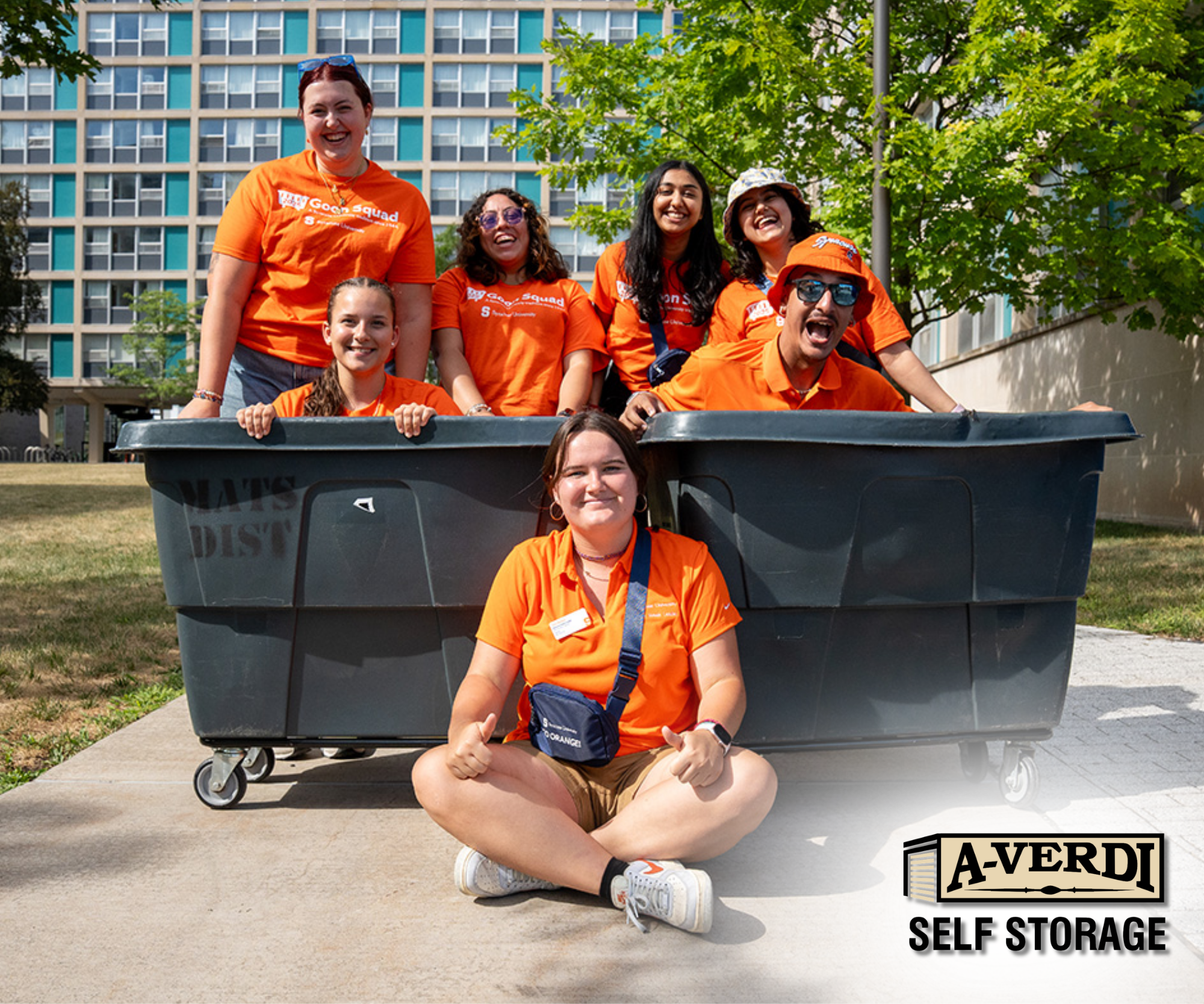 College Student Storage Made Simple: A Stress-Free Solution for Every Semester Syracuse University students in orange move-in crew shirts pose with large rolling bins outside a dorm building, with the A-Verdi Self Storage logo in the corner.