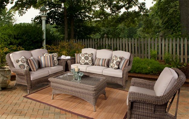 Outdoor patio seating area with wicker furniture, including a glass-top coffee table, two loveseats, and an armchair with beige cushions and decorative pillows, set on a brick patio with a rug, surrounded by greenery and a wooden fence.
