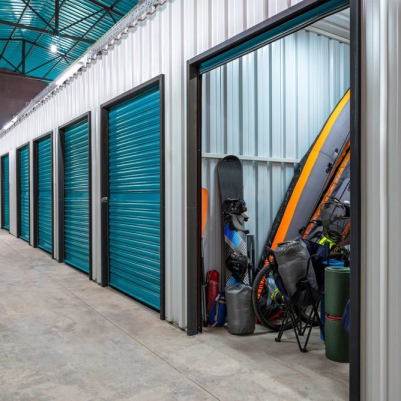 Interior of a modern self-storage facility with a row of teal roll-up doors. One unit is open, revealing stored recreational equipment including a paddleboard, kayak, snowboard, bicycle, camping chairs, sleeping bags, and other outdoor gear. The walls are made of corrugated metal, and the facility has a clean concrete floor and bright overhead lighting.