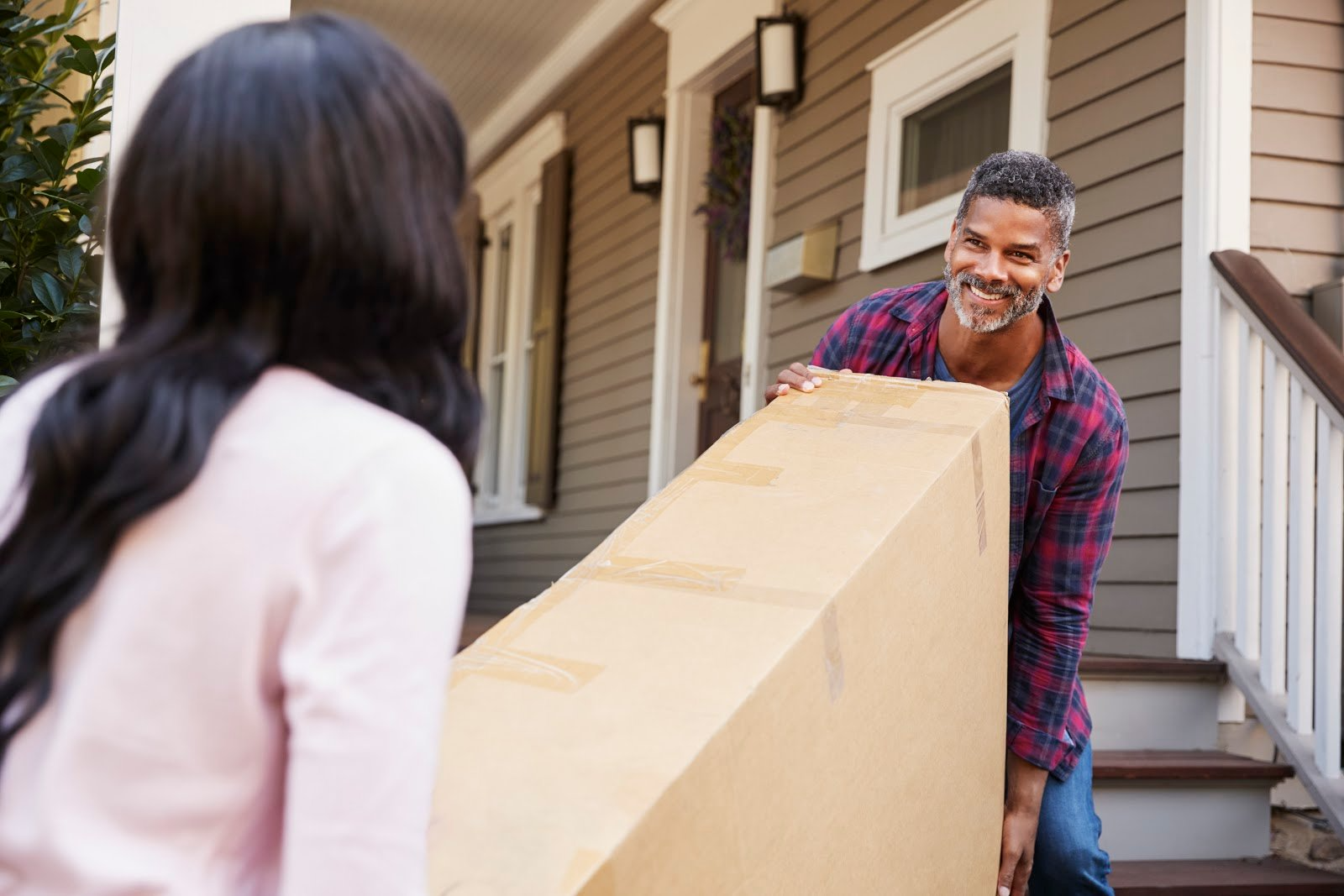 A couple carrying a large box up the stairs and into the front door of a  house.