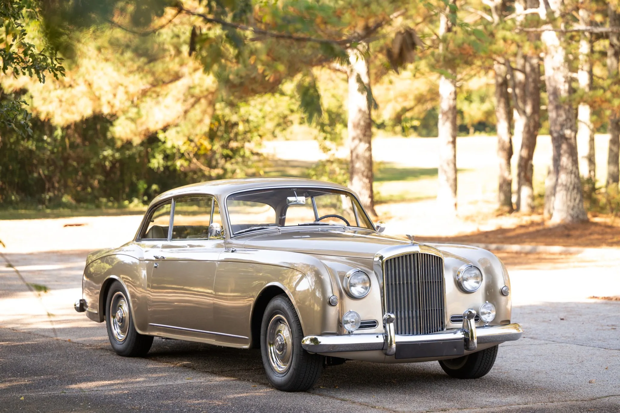 Classic silver-over-tan Bentley S2 Continental two-door saloon parked beneath trees in golden sunlight, showcasing iconic vertical grille and polished chrome details.