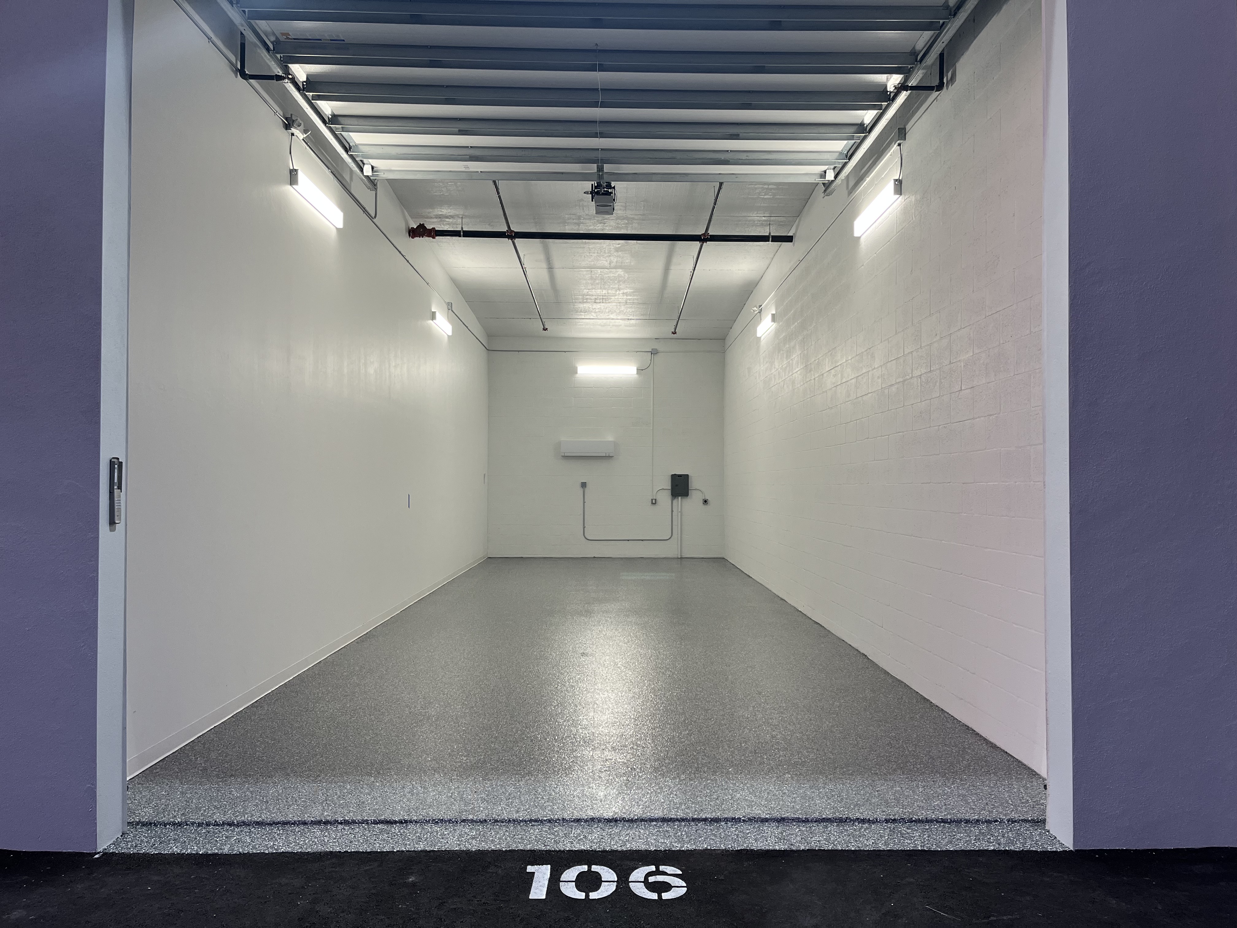 MetroMaxx Single Unit Interior - Unit 106 View into a single climate-controlled storage unit at MetroMaxx in Fort Myers, showing white walls, a polished gray epoxy floor, wall-mounted air conditioning, fluorescent lighting, and a large open roll-up door with unit number 106 painted on the ground outside.