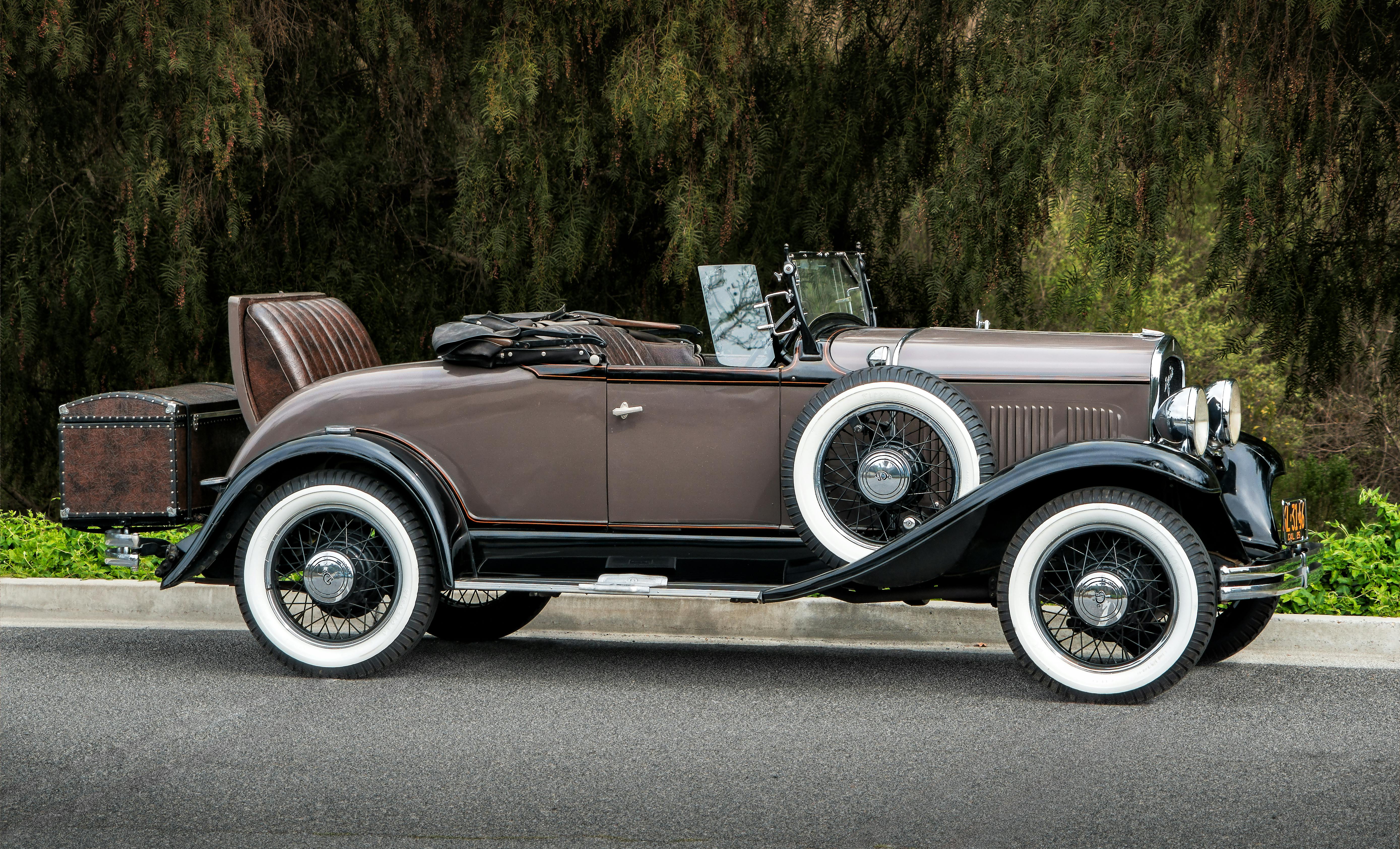 Side profile of a restored 1930s Ford Model A roadster in two-tone brown and black paint with whitewall tires, wire wheels, rumble seat, trunk-mounted luggage, and folding windshield parked outdoors