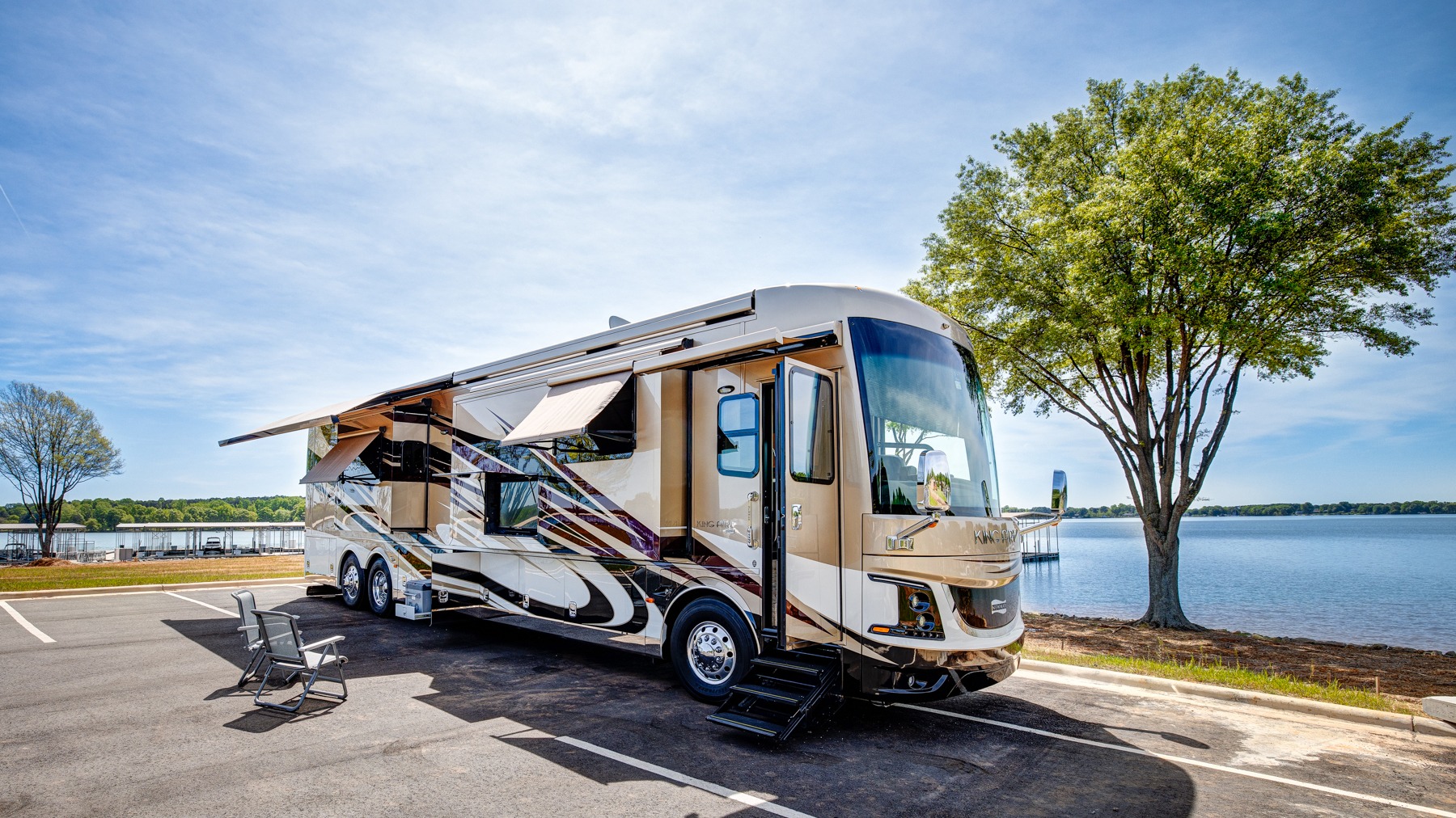 Luxury Newmar King Aire Class A motorcoach with slides and awning extended, parked lakeside on a sunny day with open entry door, steps deployed, and folding chair set up