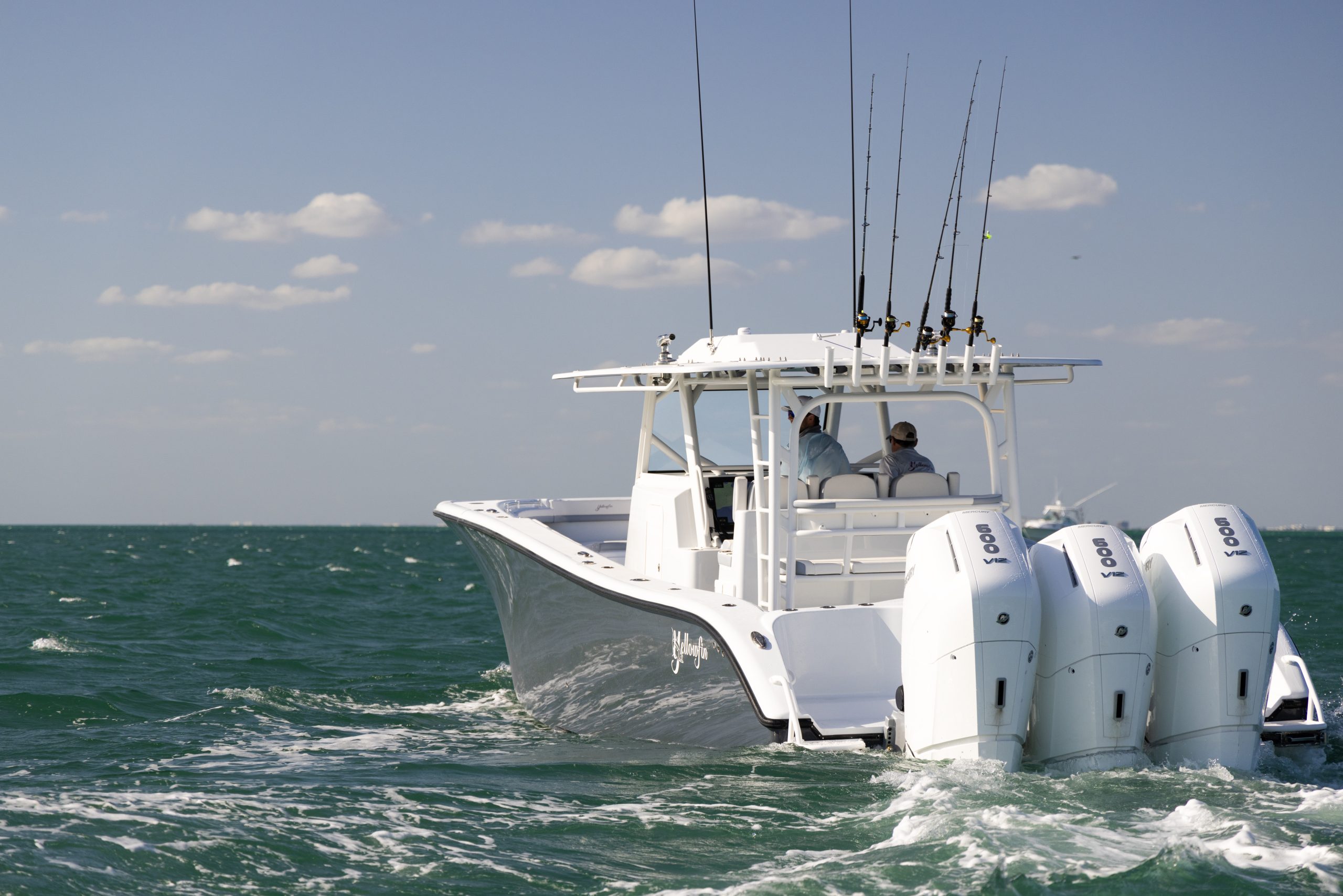 Yellowfin center console sportfishing boat with triple Yamaha 300 V8 outboard engines powering through open ocean, featuring full upper tower, outriggers, multiple mounted fishing rods, and two anglers at the helm