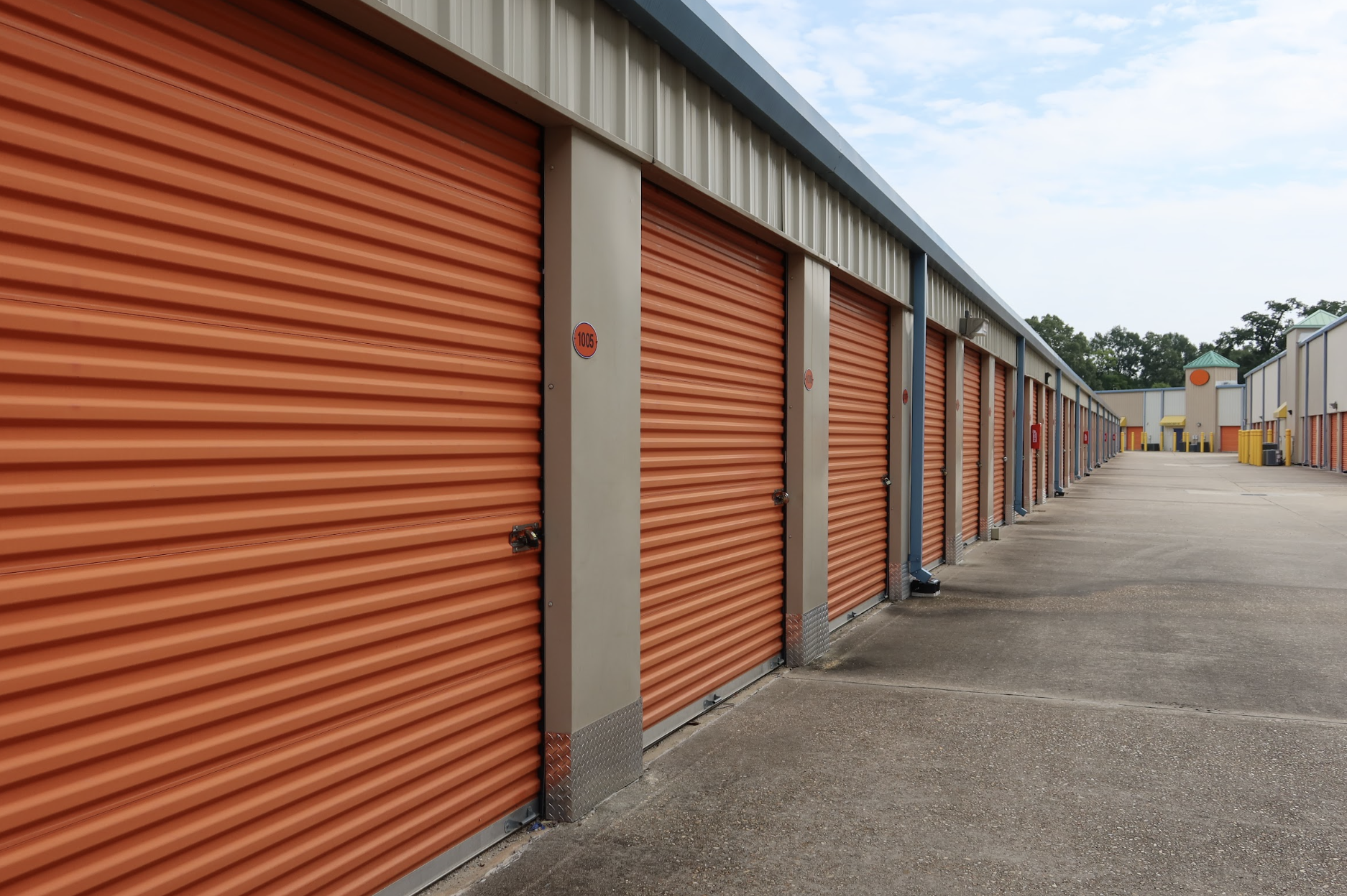 Row of drive-up storage units with orange roll-up doors along a paved driveway at Dependable Storage in Prairieville.