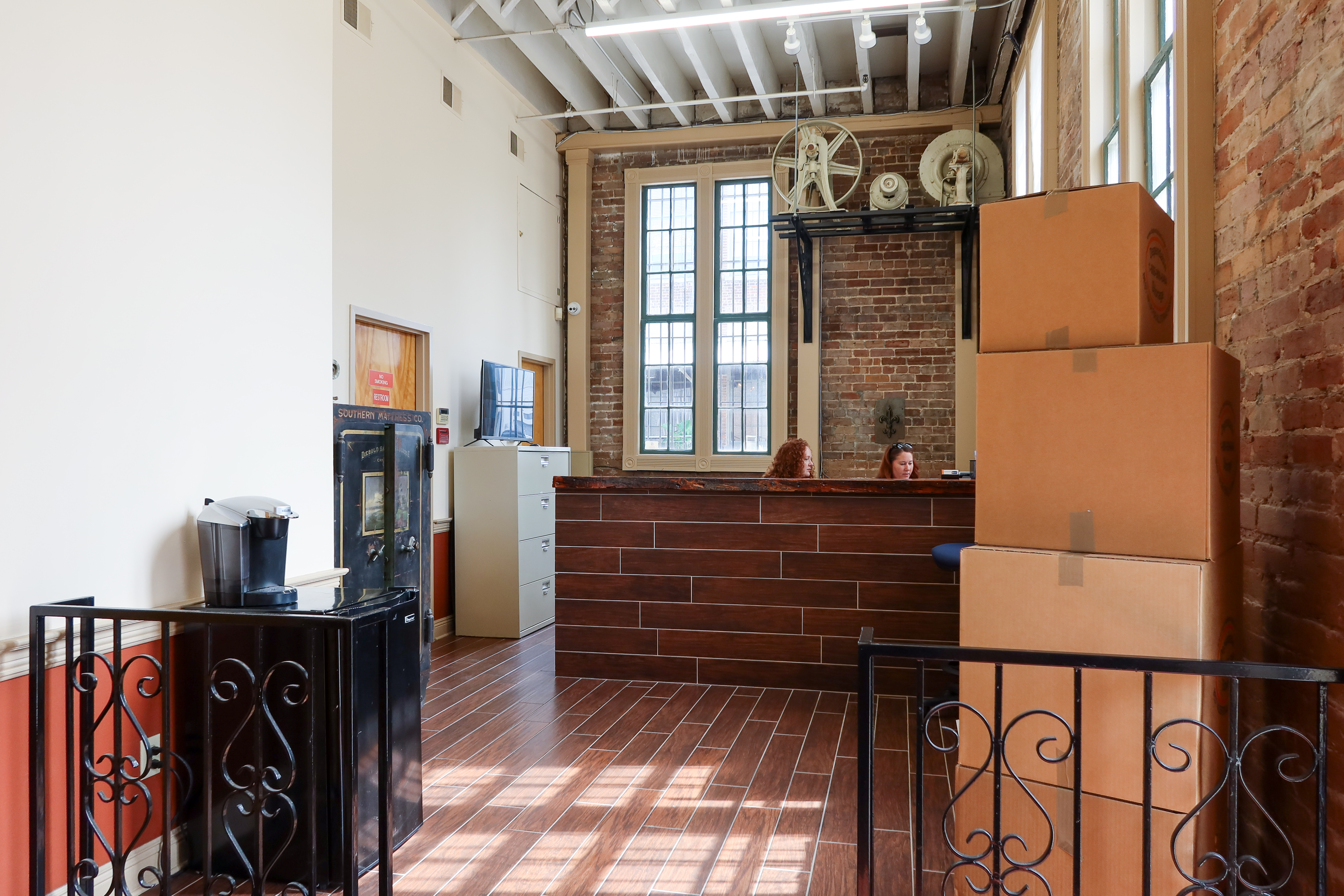 A welcoming storage facility reception area with a modern wooden counter, exposed brick walls, large windows, and neatly stacked cardboard boxes. Two staff members are seated behind the counter.