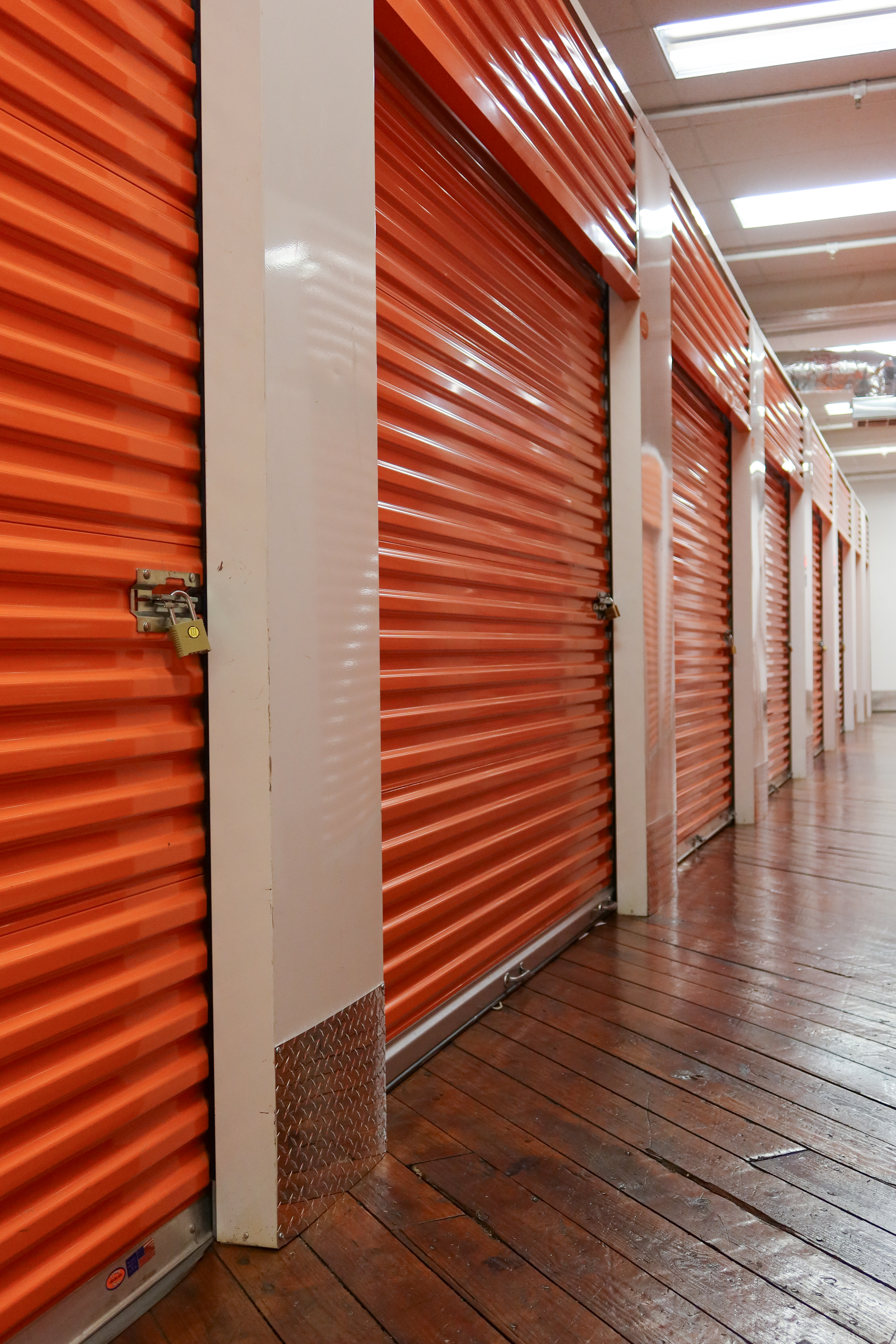 A row of orange roll-up storage unit doors with padlocks, set against a polished wooden floor and well-lit hallway.