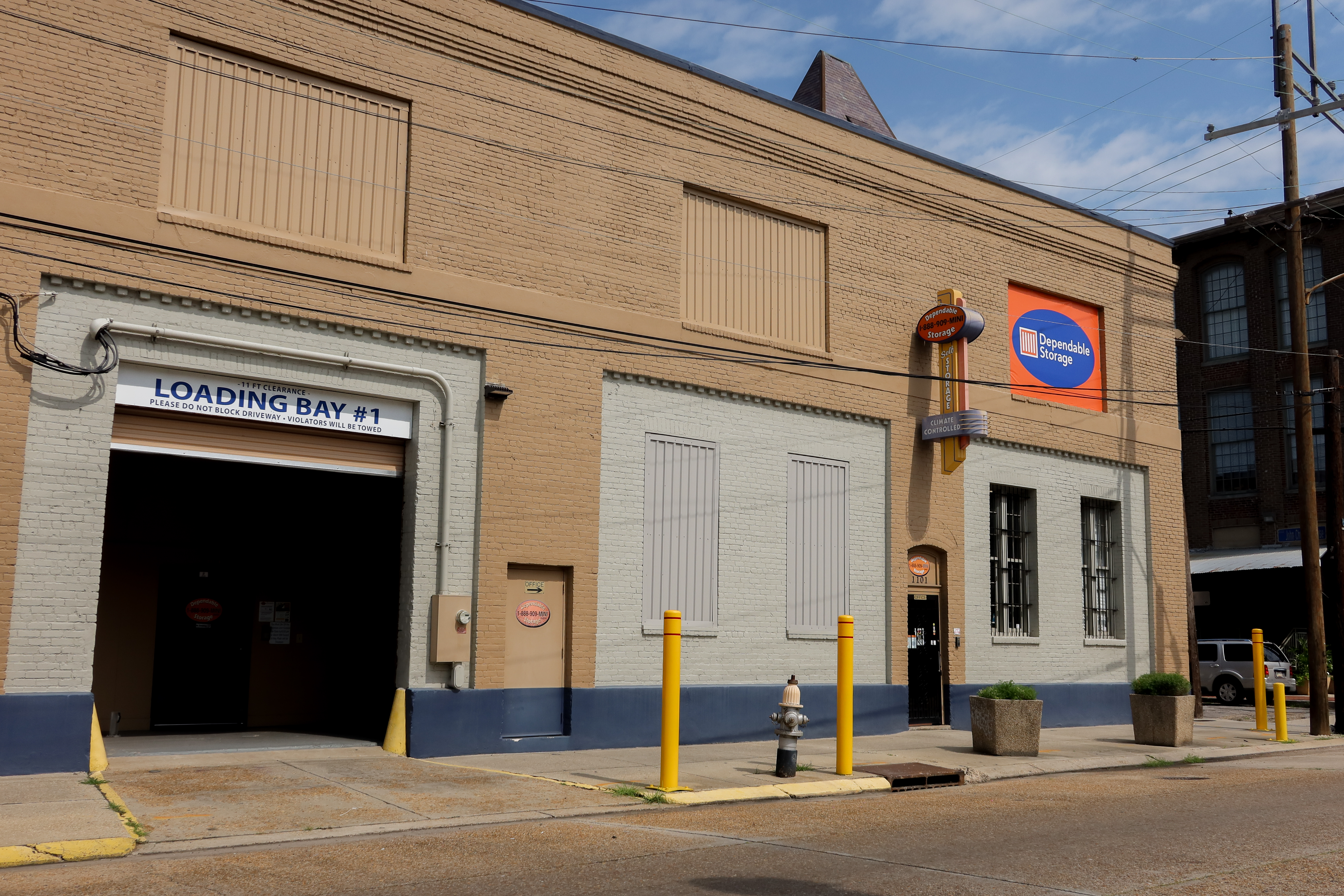 The exterior of a Dependable Storage facility featuring a large loading bay, a prominent company sign, and yellow safety bollards. The building has a beige brick facade with a blue accent at the bottom.