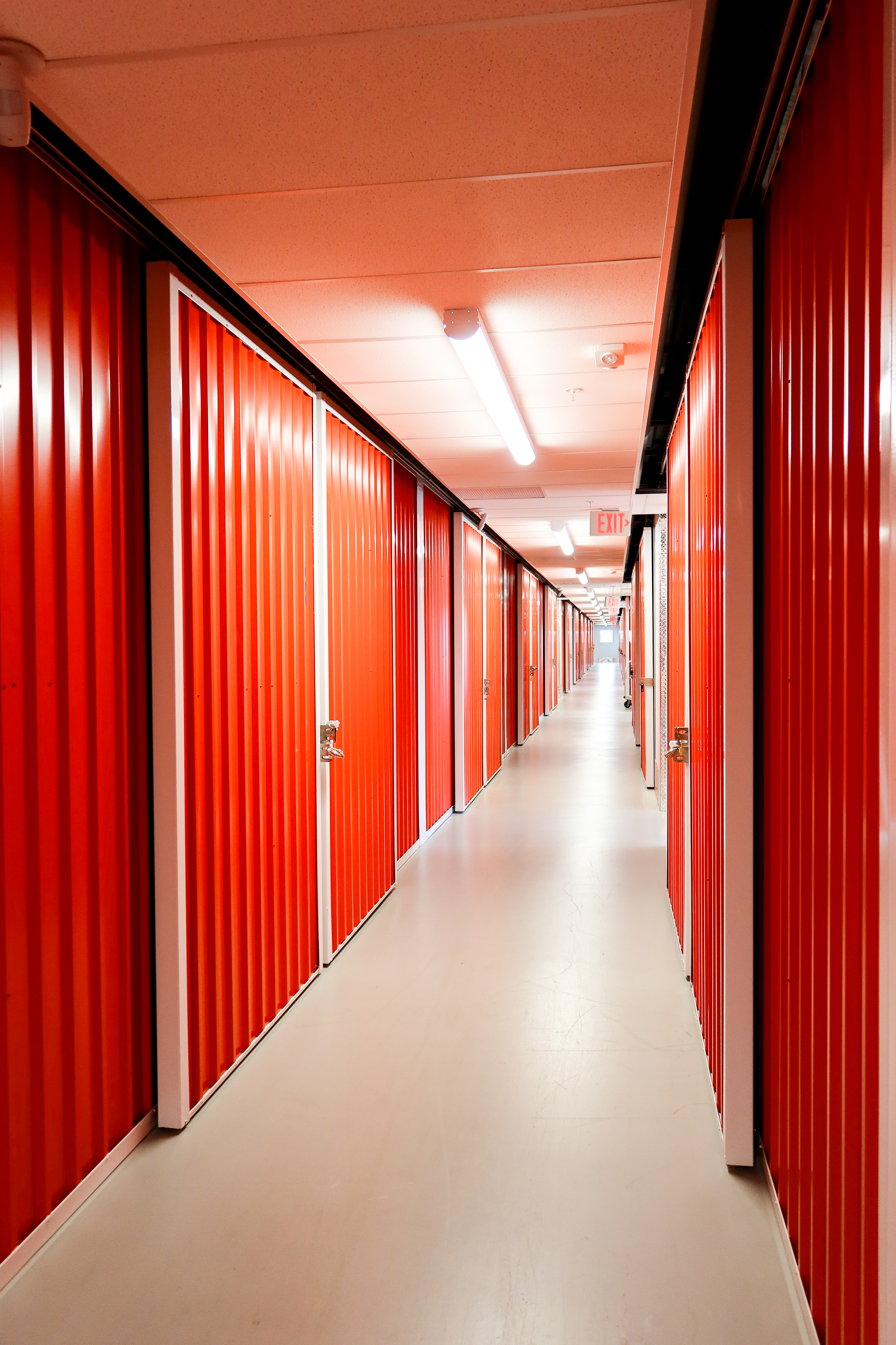 A row of orange roll-up storage unit doors with padlocks, set against a polished wooden floor and well-lit hallway.