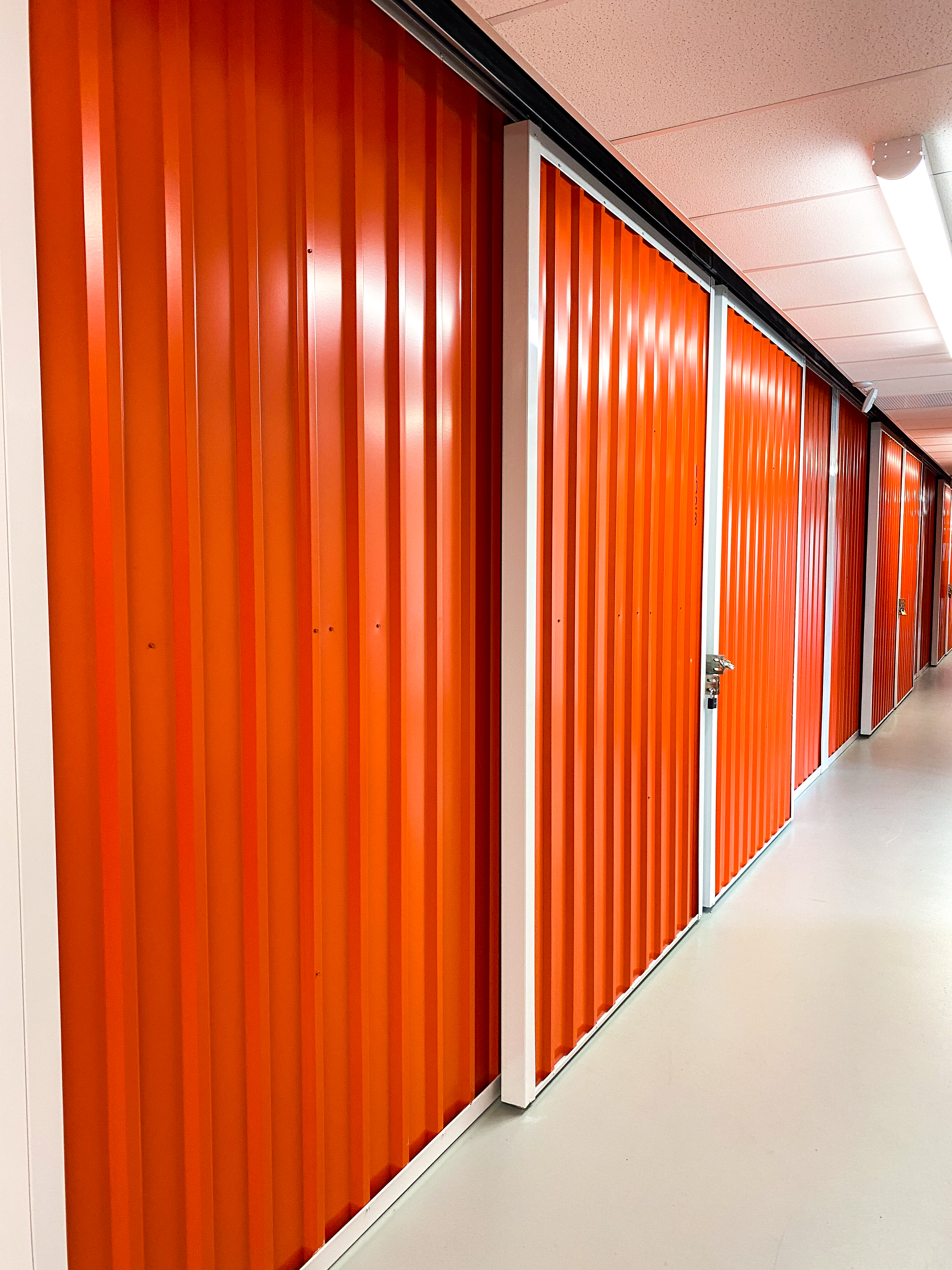A clean, well-lit hallway with orange sliding storage unit doors on both sides, leading to the back of the facility.
